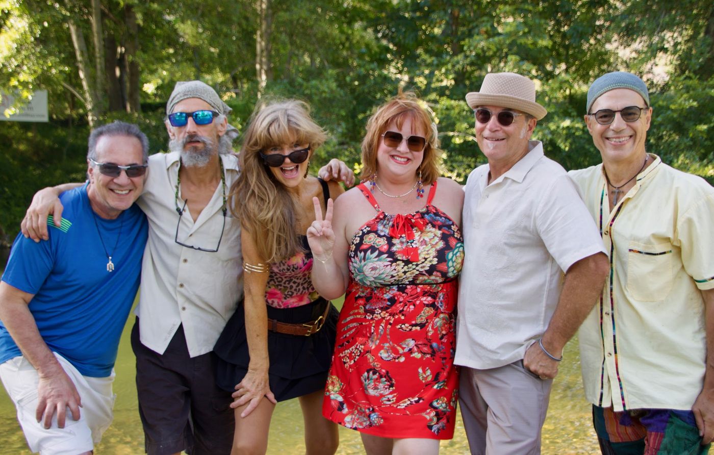 The Pozitronics standing together in a shallow creek under green trees, smiling, Kristen flashing a peace sign